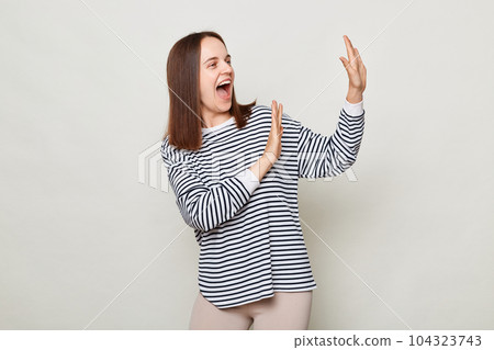 Positive optimistic brown haired woman wearing striped shirt posing isolated over gray background standing with raised palms screaming with happiness festive mood. Positive optimistic brown haired woman wearing striped shirt posing isolated over gray background standing with raised palms screaming with happiness festive mood. 104323743