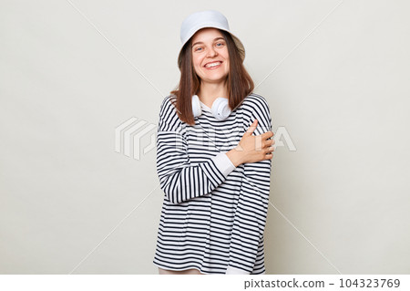 Pleased woman wearing striped shirt and panama standing with headphones over neck isolated over gray background hugging herselfexpressing self loving. Pleased woman wearing striped shirt and panama standing with headphones over neck isolated over gray background hugging herselfexpressing self loving. 104323769