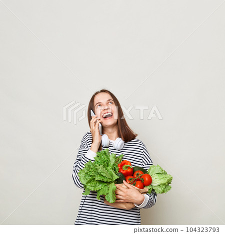 Amazed woman with brown hair wearing striped shirt standing isolated over gray background talking on mobile phone looking up at advertisement area holding vegetables 104323793