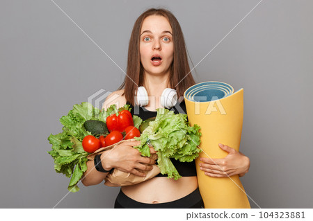 Surprised astonished Caucasian woman holding organic vegetables and yoga mat standing isolated over gray background looking at camera with big eyes and open mouth. Surprised astonished Caucasian woman holding organic vegetables and yoga mat standing isolated over gray background looking at camera with big eyes and open mouth. 104323881