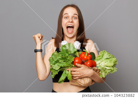 Overjoyed happy Caucasian woman holding fresh organic vegetables standing isolated over gray background clenched fist buying food in shop or market screaming with happiness. Overjoyed happy Caucasian woman holding fresh organic vegetables standing isolated over gray background clenched fist buying food in shop or market screaming with happiness. 104323882