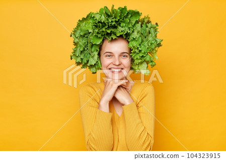 Cute smiling woman wearing casual shirt and lettuce wreath standing isolated over yellow background keeps hands under chin looking at camera with charming smile. 104323915