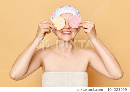 Face cleansing. Cheerful attractive young woman covering eyes with soft celluloses sponges, washing her face wearing shower cap isolated over beige background. 104323939