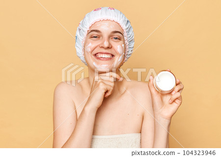Healthy skin care. Wellness through cosmetics. Anti-aging routine. Happy woman wrapping in towel with shower cap on head cream in her hands isolated over beige background. 104323946