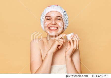 Smiling happy attractive woman in bath cap and wrapped in towel posing after taking shower applying face cream enjoying beauty procedures at home. 104323948