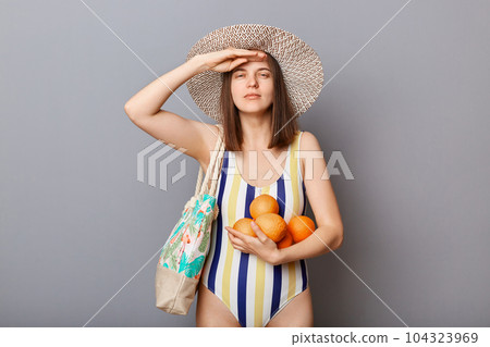 Serious woman wearing swimsuit isolated on gray background standing with oranges keeps hand near forehead looking far waiting her tropical vacation. Serious woman wearing swimsuit isolated on gray background standing with oranges keeps hand near forehead looking far waiting her tropical vacation. 104323969