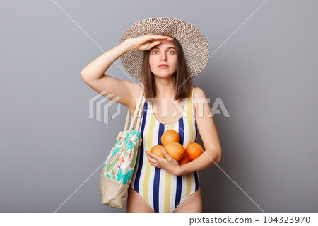 Shocked surprised astonished woman wearing striped swimsuit and straw hat isolated gray background standing with fresh oranges keeps hand near forehead looking far. Shocked surprised astonished woman wearing striped swimsuit and straw hat isolated gray background standing with fresh oranges keeps hand near forehead looking far. 104323970