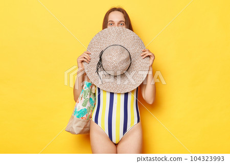 Funny woman wearing swimsuit isolated on yellow background covering half of her face with her straw hat looking with satisfied expression relaxing at exotic tour. Funny woman wearing swimsuit isolated on yellow background covering half of her face with her straw hat looking with satisfied expression relaxing at exotic tour. 104323993