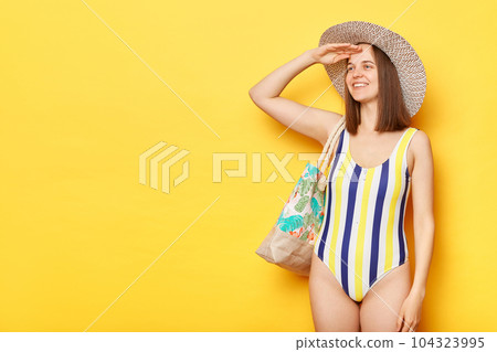 Cute smiling woman wearing swimsuit isolated on yellow background looking far with hand near forehead expressing positive emotions copy space for promotions. Cute smiling woman wearing swimsuit isolated on yellow background looking far with hand near forehead expressing positive emotions copy space for promotions. 104323995