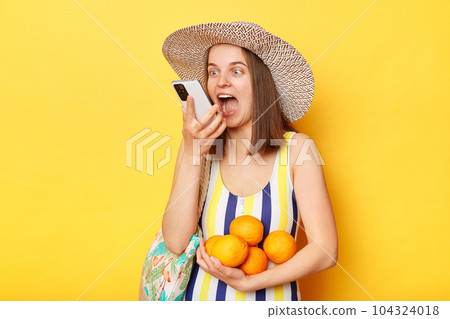 Happy woman wearing striped swimsuit and straw hat isolated yellow background using smart phone recording voice message on summer vacation. 104324018