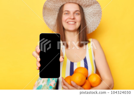 Smiling caucasian woman wearing striped swimsuit and straw hat isolated yellow background showing empty smart phone screen advertisement area holding oranges. Smiling caucasian woman wearing striped swimsuit and straw hat isolated yellow background showing empty smart phone screen advertisement area holding oranges. 104324030