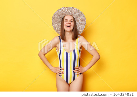Happy positive woman wearing striped swimming suit and sun hat isolated on yellow background, enjoying her holiday, keeps hands on hips, looking at camera. 104324051