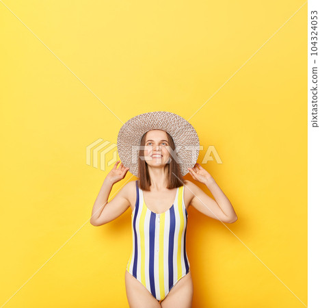 Joyful attractive woman wearing striped one-piece swimsuit and straw hat posing isolated over yellow background looking smiling upwards copy space for promotional text. 104324053