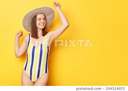 happy overjoyed woman wearing striped swimming suit and hat isolated on yellow background dancing rejoicing her vacation copy space for promotional text. happy overjoyed woman wearing striped swimming suit and hat isolated on yellow background dancing rejoicing her vacation copy space for promotional text. 104324055