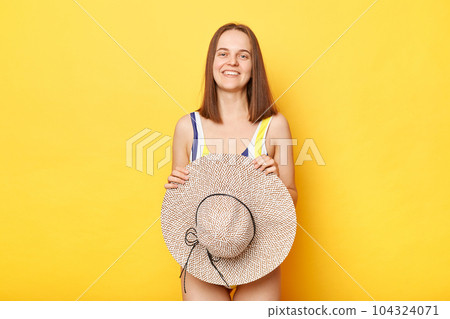 Calm pretty positive woman wearing striped swimming suit and holding hat looking at camera with smile enjoying summertime isolated on yellow background Calm pretty positive woman wearing striped swimming suit and holding hat looking at camera with smile enjoying summertime isolated on yellow background 104324071
