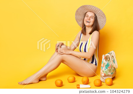 Relaxed satisfied woman wearing striped one-piece swimsuit and straw hat posing isolated over yellow background laughing to camera enjoying summertime and vacation. 104324279