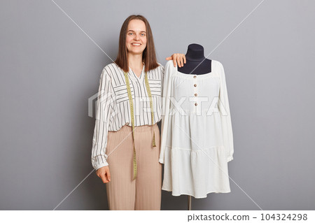 Textile production process. Successful sewing business. Professional fashion designer. Beautiful happy woman sewer standing near new dress on mannequin iisolated over gray background. 104324298