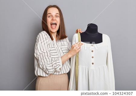 Sewing freelancer's success. Small clothing workshop. Excited amazed woman seamstress standing near new dress on mannequin iisolated over gray background. Sewing freelancer's success. Small clothing workshop. Excited amazed woman seamstress standing near new dress on mannequin iisolated over gray background. 104324307