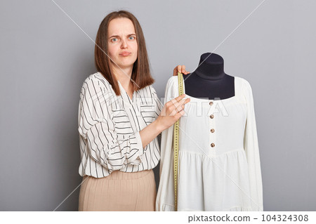 Sad dissatisfied young adult female sewer standing near mannequin with new dress isolated over gray background, looking at camera with confused expressing holding measure tape. Sad dissatisfied young adult female sewer standing near mannequin with new dress isolated over gray background, looking at camera with confused expressing holding measure tape. 104324308