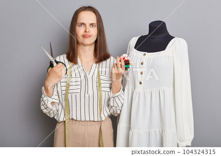 Creative atelier for fashion. Cloth and textile production. Unhappy sad woman sewer standing near new dress on mannequin iisolated over gray background. 104324315
