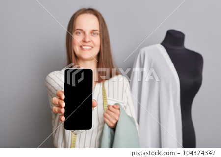 Pretty smiling professional female sewer in atelier standing near manikin and showing smart phone with empty display copy space for advertisement online ordering dressmaking service. 104324342