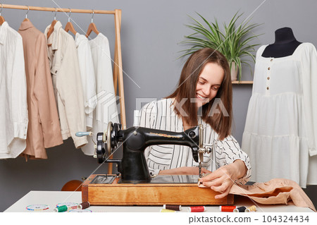 Satisfied smiling happy seamstress businesswoman working on sewing machine, sewing clothes fabric in tailor workshop, enjoying her job. Satisfied smiling happy seamstress businesswoman working on sewing machine, sewing clothes fabric in tailor workshop, enjoying her job. 104324434