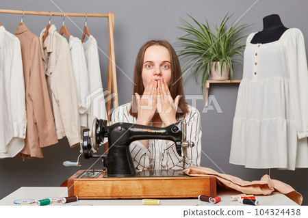 Shocked surprised woman sewer posing at her workplace sewing clothing, made mistake, dressmaker making wrong stitch, spoiled material for dress, covering mouth with hands. Shocked surprised woman sewer posing at her workplace sewing clothing, made mistake, dressmaker making wrong stitch, spoiled material for dress, covering mouth with hands. 104324438