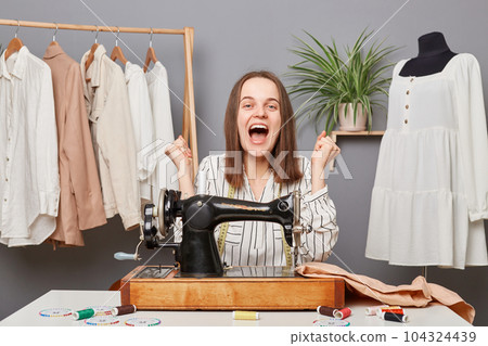 Extremely happy professional woman seamstress sewing clothing on her machine in tailor workshop, designer sewer finishing dress for new collection, clenched fists, celebrating success. Extremely happy professional woman seamstress sewing clothing on her machine in tailor workshop, designer sewer finishing dress for new collection, clenched fists, celebrating success. 104324439