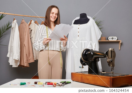 Fashion woman designer making holding papers with sketch in workshop, thinking about new collection, smiling, seamstress creating new outfits. 104324454