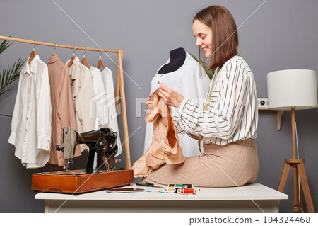 Portrait of caucasian woman in sewing workshop. holding detail of new dress, sitting on table, smiling, designer creating new collection. Portrait of caucasian woman in sewing workshop. holding detail of new dress, sitting on table, smiling, designer creating new collection. 104324468