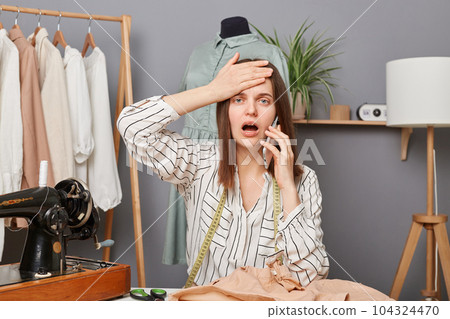 Shocked caucasian young adult female sewer working in atelier, talking on mobile phone, making facepalm gesture, having problems with supply of new fabrics. Shocked caucasian young adult female sewer working in atelier, talking on mobile phone, making facepalm gesture, having problems with supply of new fabrics. 104324470