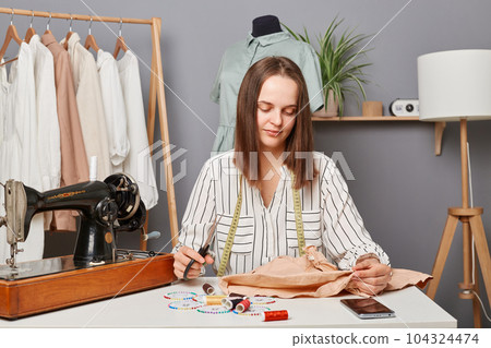 Young focused concentrated woman sewer sitting at her workplace in atelier near sewing machine with scissors in hands, thinking how to alter an old dress or shirt. Young focused concentrated woman sewer sitting at her workplace in atelier near sewing machine with scissors in hands, thinking how to alter an old dress or shirt. 104324474