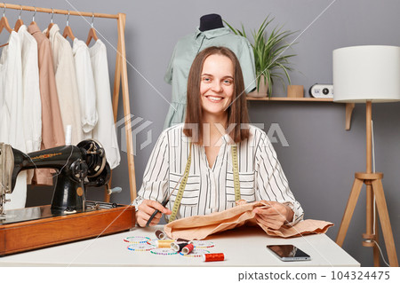 Tailoring. Woman tailors sewing fabric, sitting at her workplace holding scissors cutting textile for new dress smiling to camera enjoying her work or hobby. Tailoring. Woman tailors sewing fabric, sitting at her workplace holding scissors cutting textile for new dress smiling to camera enjoying her work or hobby. 104324475