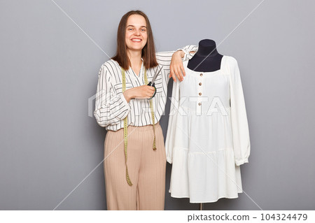 Freelance sewing success. Skilled seamstress work. Small business tailor. Smiling woman seamstress standing near new dress on mannequin iisolated over gray background. Freelance sewing success. Skilled seamstress work. Small business tailor. Smiling woman seamstress standing near new dress on mannequin iisolated over gray background. 104324479