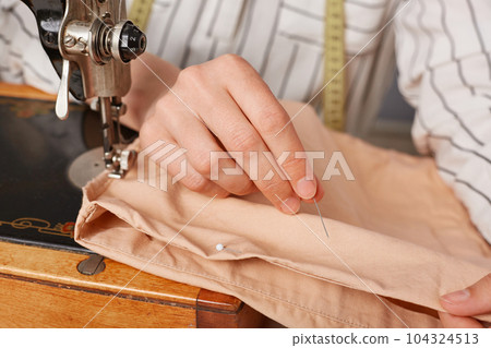 Unrecognizable seamstress sewer working on sewing machine, sewing clothes fabric in tailor workshop, making new clothing collection. Unrecognizable seamstress sewer working on sewing machine, sewing clothes fabric in tailor workshop, making new clothing collection. 104324513