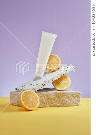 On a pastel purple background, white plastic tube displayed with marble brick, block of stone and halves of lemon. Cosmetic mockup for advertising and branding with lemon extract. Front view 104324589