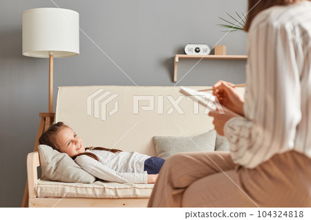 Cute relaxed little girl lying on sofa in psychological office having consultation with doctor specialist children depression stressed kid domestic violence. 104324818