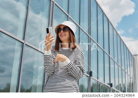 Joyful smiling woman wearing striped shirt using phone posing near building with big windows making selfie on her cell phone communicating with subscribers. Joyful smiling woman wearing striped shirt using phone posing near building with big windows making selfie on her cell phone communicating with subscribers. 104324964