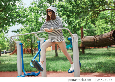 Happy smiling girl doing ellipsoid on outdoor sport ground woman training on ellipsoid while walking with beautiful green nature on background. Happy smiling girl doing ellipsoid on outdoor sport ground woman training on ellipsoid while walking with beautiful green nature on background. 104324977