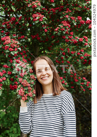 Portrait of young adult charming pretty woman wearing striped shirt posing near tree blossoms blooming flowers in garden park in summer nature. Portrait of young adult charming pretty woman wearing striped shirt posing near tree blossoms blooming flowers in garden park in summer nature. 104324979