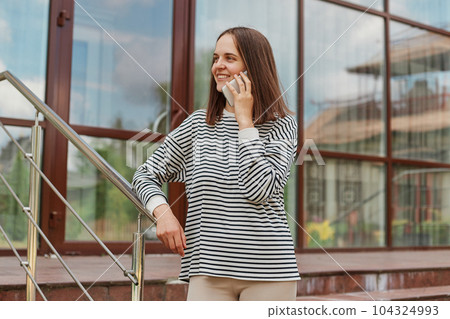 Dark haired adult woman talking on her phone against building with big windows, smiling, having pleasant conversation while walking in town 104324993