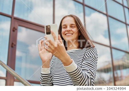 Joyful smiling woman wearing striped shirt using phone for making video call posing near building with big windows holding her cell phone communicating waving hand with hand Joyful smiling woman wearing striped shirt using phone for making video call posing near building with big windows holding her cell phone communicating waving hand with hand 104324994