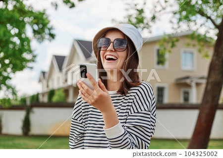 Laughing positive woman posing on city street with smartphone on background laughing with happy expression using mobile phone for making selfie. Laughing positive woman posing on city street with smartphone on background laughing with happy expression using mobile phone for making selfie. 104325002