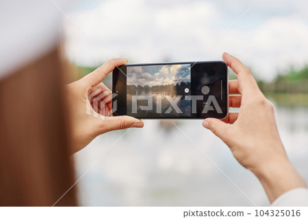 Unrecognizable female blogger using cellular technology for shooting video content during travel vacations, woman with smartphone device photographing lake or river. 104325016
