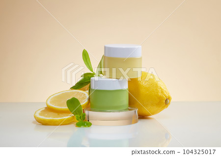 Two blank label jars in green and yellow color stacked on transparent podium. Displayed with lemon and green leaves. Natural cosmetic concept with Lemon (Citrus limon) 104325017