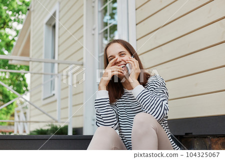 Dark haired adult woman wearing striped shirt and leggins sitting on house porch talking on mobile phone smiling happily having conversation with friends. 104325067