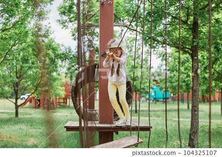 Full length portrait of little girl in cap playing on rope swings at sunny playground having fun alone in sunny day overcomes obstacles. Full length portrait of little girl in cap playing on rope swings at sunny playground having fun alone in sunny day overcomes obstacles. 104325107