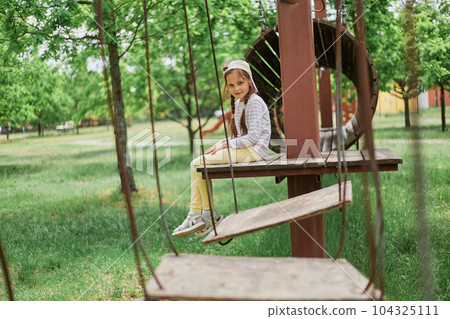 Little tired girl wearing casual attire and cap sitting on rope swings at sunny playground having fun resting having break of active playing. 104325111