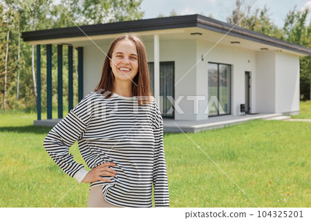 Dark haired smiling standing near building new house on green lawn keeps hand on hip smiling to camera wearing striped shirt having positive emotions. 104325201