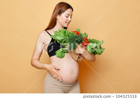 Beautiful pregnant woman with bare belly embraces and looking at fresh vegetables posing isolated over beige background healthy eating during pregnancy. Beautiful pregnant woman with bare belly embraces and looking at fresh vegetables posing isolated over beige background healthy eating during pregnancy. 104325318